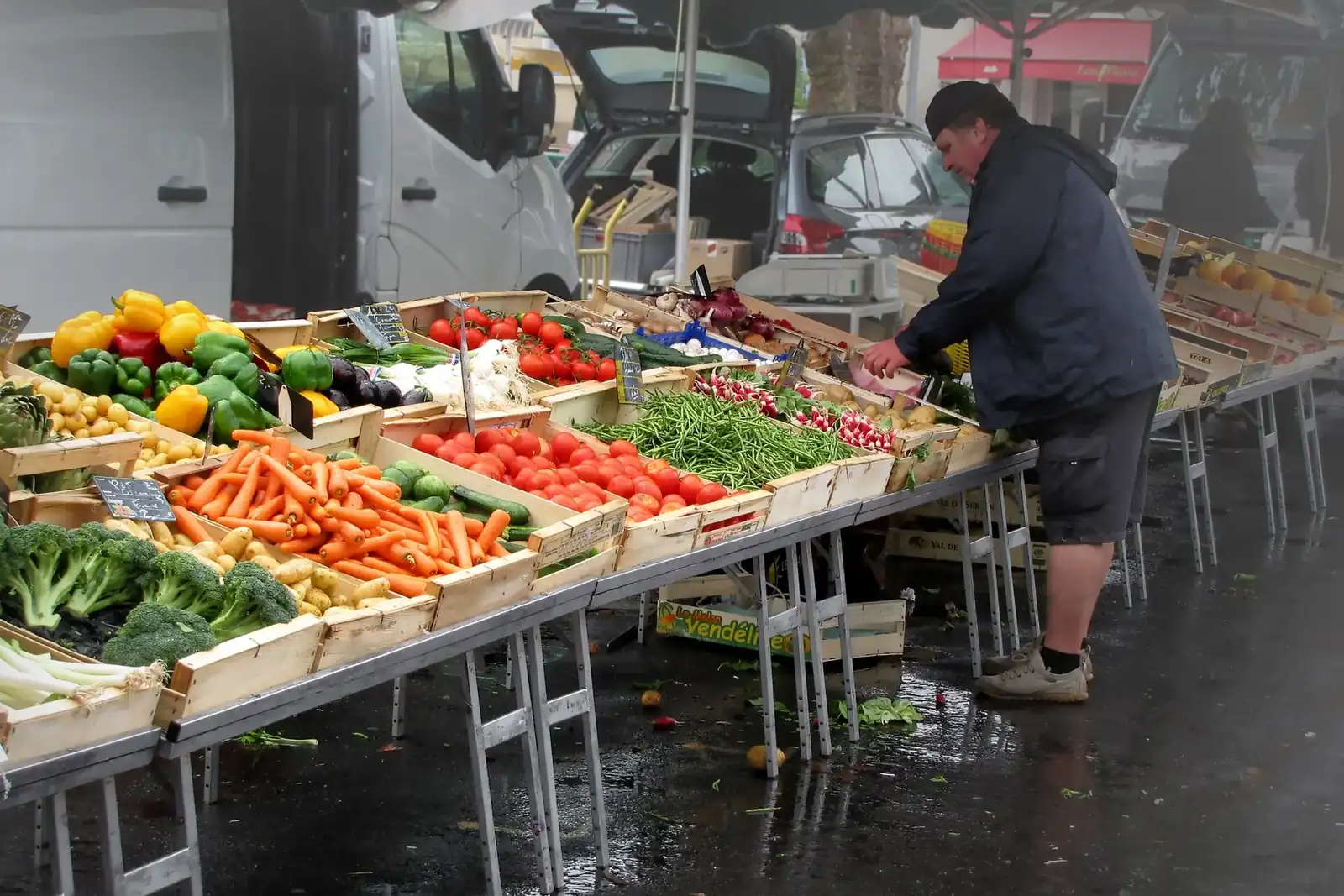 Un étal de primeur sur un marché de plein air, avec des cagettes de tomates, de carottes, de courgettes et de salades sous une bâche
